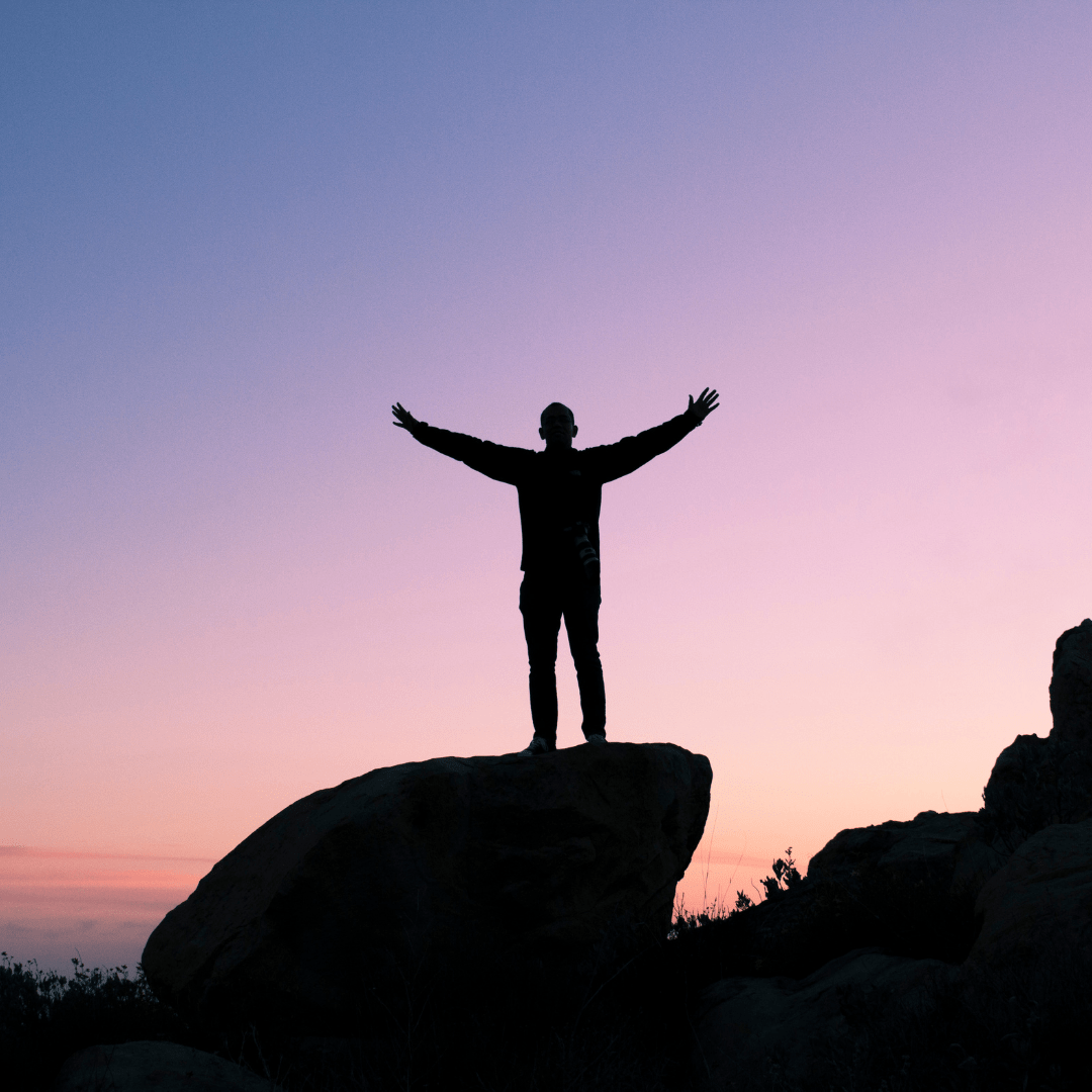 Man standing on top of a large flat rock in triumph gazing out over a sunset in the valley below